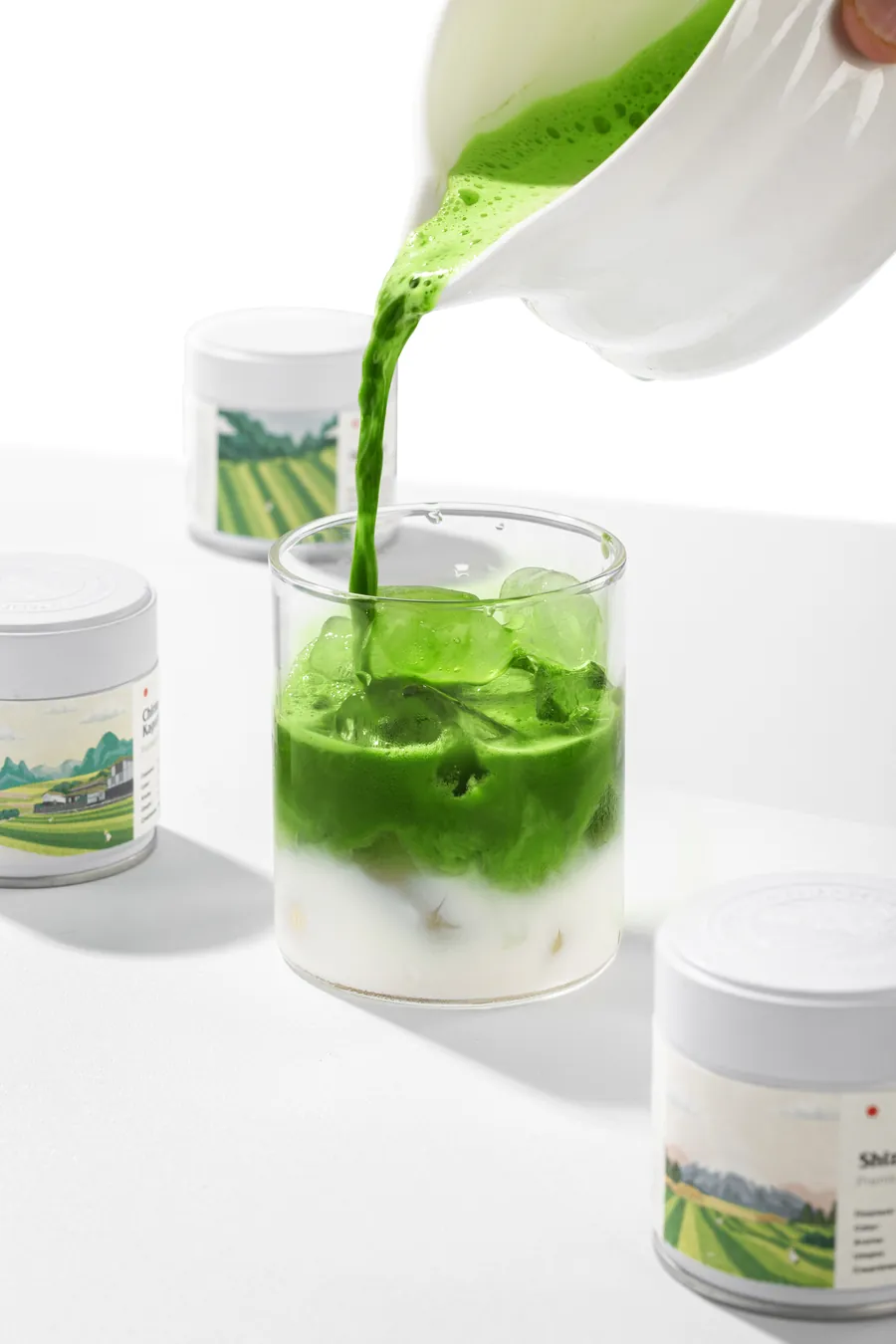 Whisked matcha poured over ice and milk in a glass, M'atelier tins behind